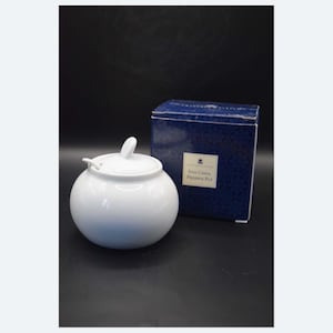 May include: A white, round porcelain sugar bowl with a lid and a small handle. The bowl is next to a blue box with the text "Fine China Preserve Pot". The background is black.