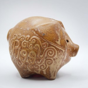 Może przedstawiać: Ceramiczna skarbonka w kolorze brązowym, o teksturowanej powierzchni i dekoracyjnym wzorze w kształcie wiru. Na boku wygrawerowano napis "PIGS". Skarbonka ma otwór na monety i mały otwór na nos.