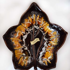 Può includere: Un piatto in ceramica a forma di foglia con finitura marrone lucido. Il piatto presenta un design a foglia stilizzato con accenti gialli e bianchi. Il piatto ha un'etichetta con la scritta "Vallauris".