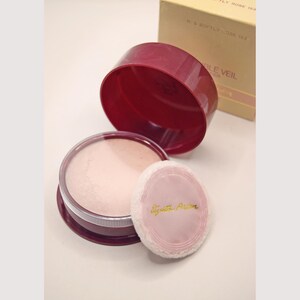 May include: A vintage makeup set with a burgundy powder compact, a powder puff, and a box. The compact is open, showing the powder. The puff is pink with gold text. The box reads "N.5 Softly Rose 153".