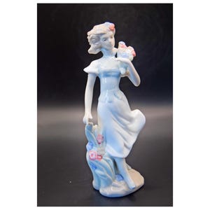 May include: A porcelain figurine of a woman in a flowing blue dress, holding a bouquet of flowers. The figurine has a light blue and white color scheme with pink and blue floral accents. The woman is standing on a small base with additional floral details.