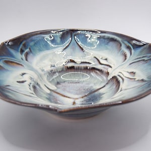 May include: A ceramic bowl with a unique, organic shape. The bowl features a blend of blue, white, and brown glaze, creating a marbled effect. The rim has a scalloped edge, adding to its artistic appeal. The bowl is suitable for serving or display.