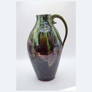 Op de afbeelding: Een hoge, geglazuurde keramische kan met een donkerbruine basis en een groen-bruin bovenstuk. De kan heeft een gebogen handvat en een brede opening. De glazuur heeft een glanzende afwerking, wat het een ambachtelijke uitstraling geeft.