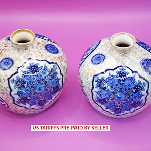 Op de afbeelding: Twee ronde keramische vazen met een witte basis, versierd met blauwe en gouden bloempatronen. Elke vaas heeft een centraal blauw ontwerp en een gouden rand. De tekst "US TARIFFS PRE-PAID BY SELLER" is zichtbaar aan de onderkant.