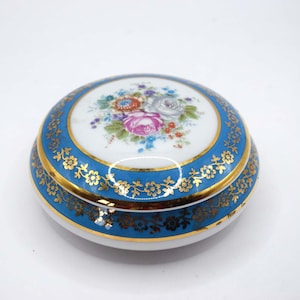 Puede incluir: Una caja de baratijas redonda de cerámica con base blanca y borde azul. La tapa presenta un diseño floral en el centro y detalles dorados. La caja mide aproximadamente 10 cm de diámetro.