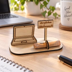 Könnte beinhalten: Ein heller Holzaufsteller mit einer lasergeschnittenen Laptop-Form und einem kleinen Schild mit der Aufschrift "Herzlichen Glückwunsch". Ein zusammengerollter Euro-Geldschein wird von einem Holzring auf der Basis gehalten.