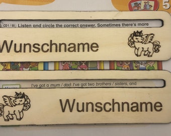 May include: Two wooden name tags featuring a unicorn design and the text "Wunschname" on each. The top tag has the text "CD1/55 Listen and circle the correct answer. Sometimes there's more" above the unicorn. The bottom tag has the text "I've got a mum/dad. I've got two brothers/sisters, and" above the unicorn.