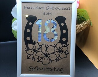 Puede incluir: Un marco de fotos blanco con una tarjeta marrón en su interior. La tarjeta tiene una herradura negra con el número 18 en su interior. La tarjeta también tiene el texto "Herzlichen Glückwunsch zum Geburtstag".