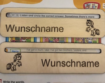 May include: Two wooden nameplates with the German word "Wunschname" engraved on them. Each nameplate has a slot for a card with a unicorn illustration and text. The top nameplate has the text "CD1/55 Listen and circle the correct answer. Sometimes there's more". The bottom nameplate has the text "I've got a mum/dad. I've got two brothers/sisters, and".