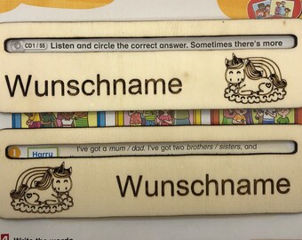 May include: Two wooden name tags featuring a unicorn and rainbow design. The text on the tags reads "Wunschname" and "CD1/55 Listen and circle the correct answer. Sometimes there's more". The text on the bottom tag reads "I've got a mum/dad. I've got two brothers/sisters, and".