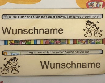 May include: Two wooden name tags with a unicorn design and the text "Wunschname" on each. The top tag has the text "CD1/55 Listen and circle the correct answer. Sometimes there's more" on it. The bottom tag has the text "I've got a mum/dad. I've got two brothers/sisters, and" on it.