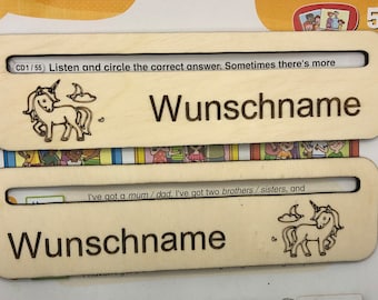 May include: Two wooden name tags with a unicorn design and the text "Wunschname" on each. The text "CD1/55 Listen and circle the correct answer. Sometimes there's more" is at the top of the first tag. The text "I've got a mum/dad. I've got two brothers/sisters, and" is at the top of the second tag.