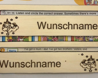 May include: Two wooden name tags with the word "Wunschname" in a dark brown font. Each tag has a slot at the top. One tag has a sleeping unicorn and stars. The text "Listen and circle the correct answer" is visible.
