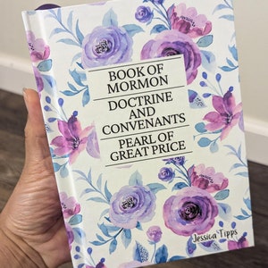 May include: A white book with a floral design featuring purple and blue flowers. The title of the book is "Book of Mormon Doctrine and Covenants Pearl of Great Price" by Jessica Tipps.
