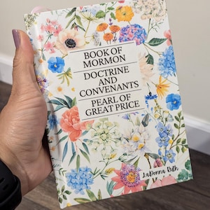 May include: A book titled "Book of Mormon, Doctrine and Covenants, Pearl of Great Price" with a floral cover design. The cover features a variety of colorful flowers, including pink, blue, and orange blooms, set against a white background.