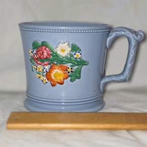 Puede incluir: Una taza de cerámica azul claro con un diseño floral. La taza presenta un relieve en relieve de flores coloridas y hojas verdes. El asa es curva y detallada. La taza mide aproximadamente 10 cm de altura.