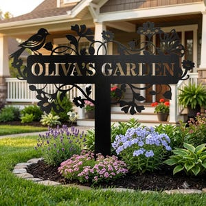 May include: A black metal garden sign featuring the text OLIVA'S GARDEN, decorated with laser-cut silhouettes of a bird, flowers, and vines. The sign is mounted on a central stake and positioned in a garden bed with various green plants.