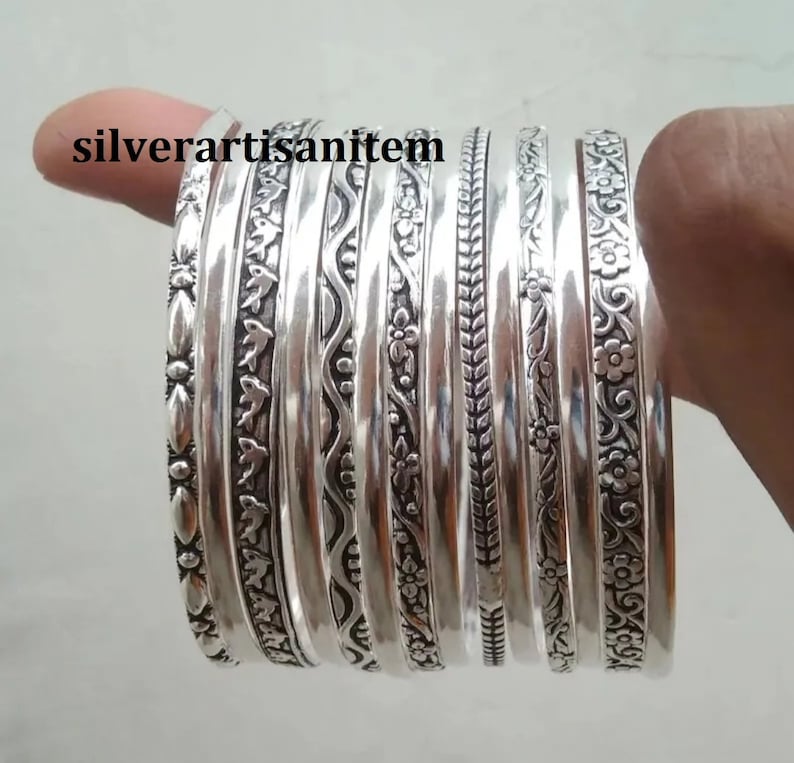 K&ouml;nnte beinhalten: Ein Set aus neun silbernen Armreifen mit aufwendigen Pr&auml;gemustern. Die Armreifen sind &uuml;bereinander gestapelt und haben eine Vielzahl von Mustern, darunter florale, geometrische und abstrakte Designs.