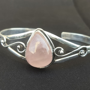 Puede incluir: Brazalete de plata con una piedra preciosa de cuarzo rosa en forma de pera. El brazalete tiene adornos en espiral y está sobre un fondo oscuro. La piedra preciosa mide aproximadamente 2,5 cm de largo.