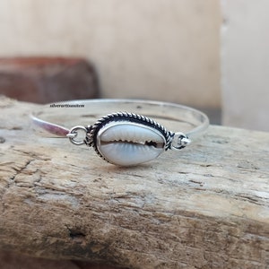 Op de afbeelding: Een zilveren armband met een witte schelpbedel. De armband heeft een gedraaid touwontwerp.