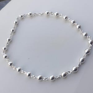 Op de afbeelding: Een delicate zilveren ketting met een reeks kleine, ronde kralen die aan elkaar zijn geregen. De kralen zijn gelijkmatig verdeeld en verbonden door een fijne ketting, waardoor een eenvoudig maar elegant ontwerp ontstaat. De ketting is waarschijnlijk gemaakt van sterling zilver.