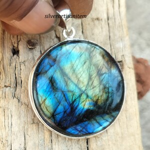 May include: Round pendant with a vibrant, iridescent labradorite stone. The stone displays shades of blue, green, and black, set in a polished silver bezel. The pendant has a silver loop for a chain.