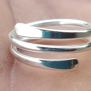 May include: A close-up of a silver ring with a spiral design. The ring has a smooth, polished surface and appears to be made of metal. The ring is resting on a skin-toned surface.