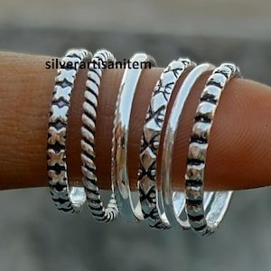 May include: Five silver stacking rings with different textured patterns. The rings are worn on a finger.