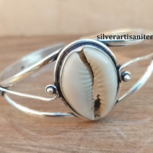 Op de afbeelding: Een zilverkleurige manchetarmband met een groot, crèmekleurig zeeschelp-middelpunt. De armband heeft een open ontwerp met sierlijke, gebogen banden en kleine decoratieve accenten. De zeeschelp is het middelpunt en toont zijn natuurlijke textuur en kleur.