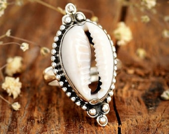 May include: A silver ring featuring a large, white cowrie shell as its centerpiece. The shell is set within a decorative, oval-shaped frame with a beaded border. The ring's design has a bohemian or coastal aesthetic.