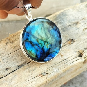 May include: A round pendant with a vibrant blue and green iridescent stone set in a silver-colored metal. The stone's surface displays a unique pattern of colors. The pendant is suspended from a silver bail.