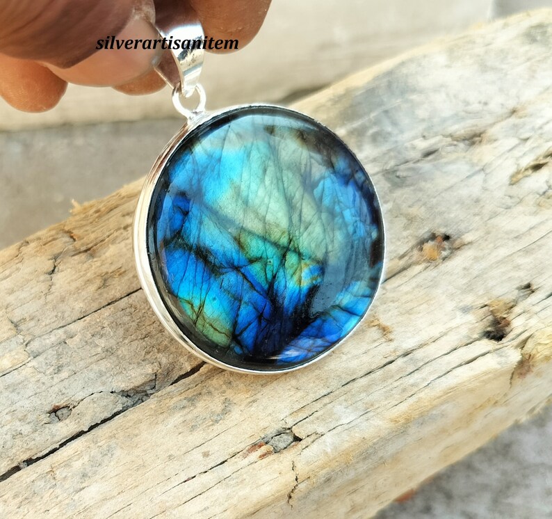 May include: A round pendant with a vibrant blue and green iridescent stone set in a silver-colored metal. The stone's surface displays a unique pattern of colors. The pendant is suspended from a silver bail.