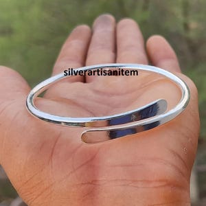 Op de afbeelding: Een zilverkleurige manchetarmband met een open ontwerp. De armband wordt in een hand gehouden en de tekst "silverartisanitem" is zichtbaar. De armband heeft een glad, reflecterend oppervlak.