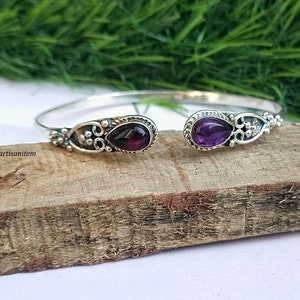 Può includere: Un braccialetto rigido in argento con due pietre preziose di ametista. Il braccialetto ha un design delicato e decorato con piccole perline e accenti floreali.