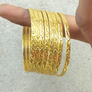Pode incluir: Um conjunto de sete pulseiras de metal dourado com um padrão floral em relevo contínuo. Cada pulseira tem uma superfície polida e reflexiva com um design repetitivo de pequenas flores e folhas ao longo de toda a circunferência.