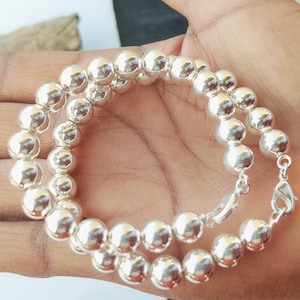 Puede incluir: Un brazalete de cuentas plateadas con un diseño de doble hebra. El brazalete presenta múltiples cuentas redondas y brillantes de plata ensartadas. El cierre también es plateado. El brazalete se muestra en la mano de una persona.