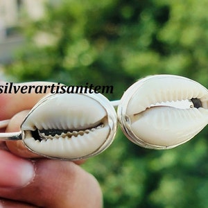 Op de afbeelding: Een zilveren armband met twee witte kauri schelpen. De schelpen zijn ingelegd in zilver en de armband is verstelbaar.
