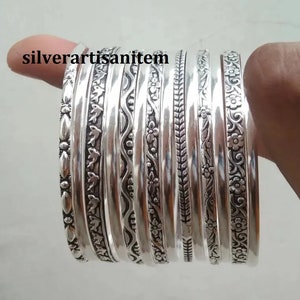 K&ouml;nnte beinhalten: Ein Set aus neun silbernen Armreifen mit aufwendigen Pr&auml;gemustern. Die Armreifen sind &uuml;bereinander gestapelt und haben eine Vielzahl von Mustern, darunter florale, geometrische und abstrakte Designs.