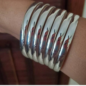 May include: A stack of silver bangles, worn on a person's wrist. The bangles are smooth and shiny, and they are stacked on top of each other.