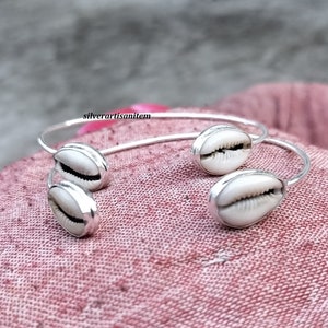May include: Two silver bangles with cowrie shell accents. The bangles are open and have a simple design.