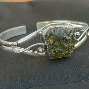 以下が含まれることがあります： 大きな緑色の粗削りされた宝石が付いたシルバーブレスレット。ブレスレットは繊細で渦巻くデザインです。