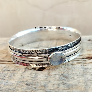 May include: A silver bangle bracelet with a moonstone gemstone. The bracelet has a hammered finish and a floral design.