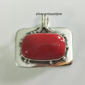 May include: A silver pendant featuring a red coral stone set within a rectangular frame. The pendant has a bail for attaching to a chain.