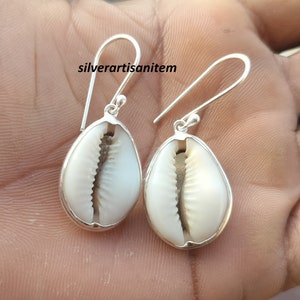 Könnte beinhalten: Ein Paar silberne Ohrringe mit einem weißen Kaurischalen-Design. Die Ohrringe haben einen einfachen Hakenverschluss.