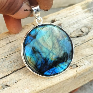 May include: Round pendant with a polished labradorite stone, showcasing iridescent blues, greens, and blacks. The stone is set in a silver bezel with a matching bail for a necklace. The pendant is held up by a hand.
