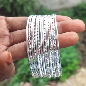 May include: A stack of silver bangles with a twisted rope design. The bangles are held in a hand, with a blurred green background. The bangles are made of a shiny, reflective metal.