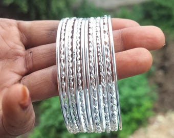 May include: A collection of silver bangles with a twisted rope pattern. The bangles are held in a hand, with a blurred green backdrop. The bangles are made of a shiny, reflective metal.