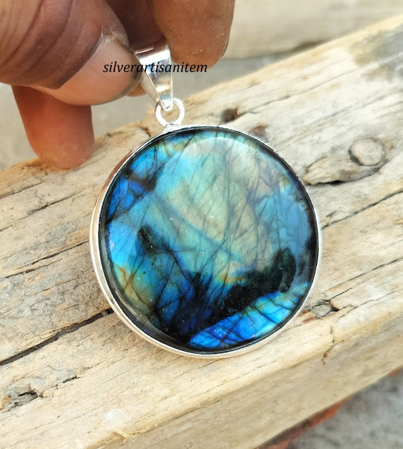 May include: Round pendant with a polished labradorite stone, showcasing iridescent blues, greens, and blacks. The stone is set in a silver bezel with a matching bail for a necklace. The pendant is held up by a hand.