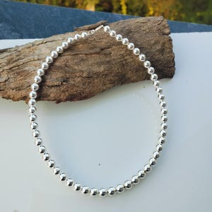 Puede incluir: Un collar de cuentas plateadas con un diseño sencillo. El collar está compuesto por pequeñas cuentas redondas y brillantes. El collar se muestra sobre un trozo de madera sobre un fondo blanco.