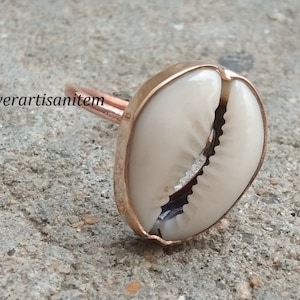 May include: A gold-toned ring with a white cowrie shell set in the center. The ring is made of metal and has a smooth finish.